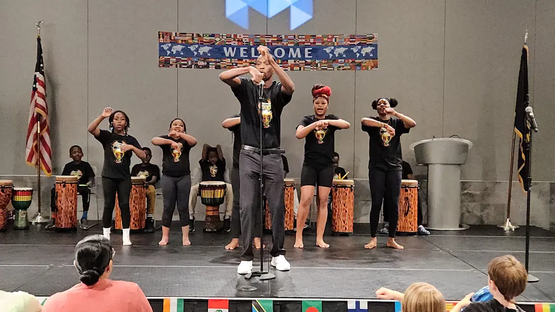 An Academy leader guides a call-and-response movement at the microphone as dancers follow along and youth drummers play behind them