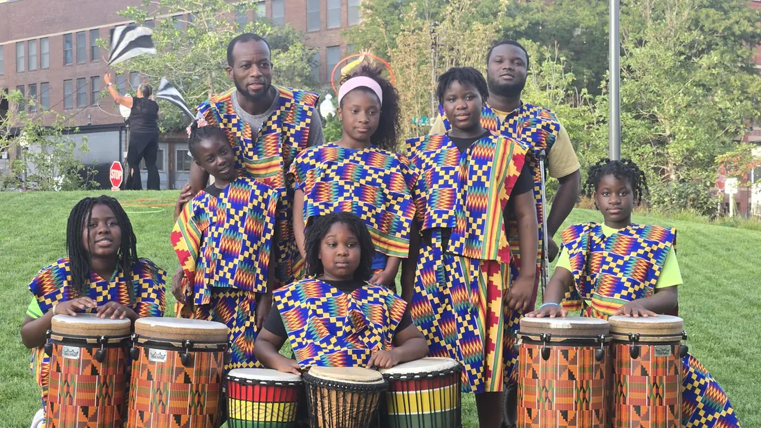 Academy youth and two instructors in matching kente-print outfits gathered behind djembe and conga-style drums on a lawn
