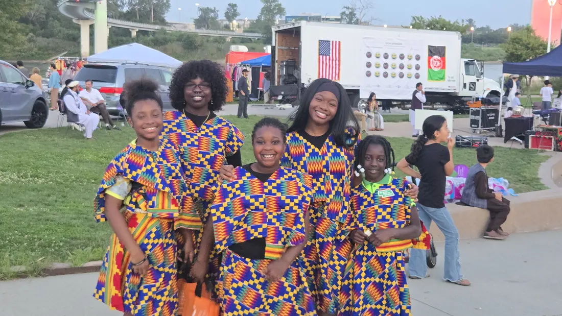 Five smiling Academy girls in matching kente-print outfits at an outdoor multicultural festival