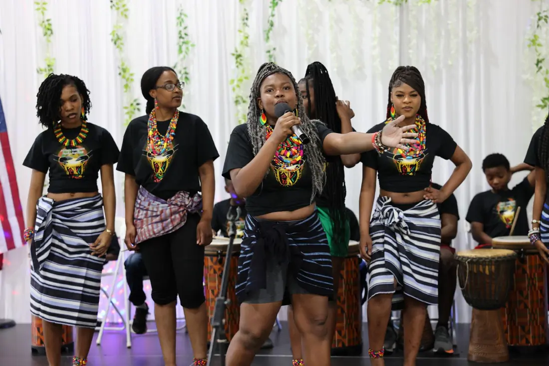 An Academy vocalist with silver braids leads a song, arm outstretched, while dancers and drummers perform behind her