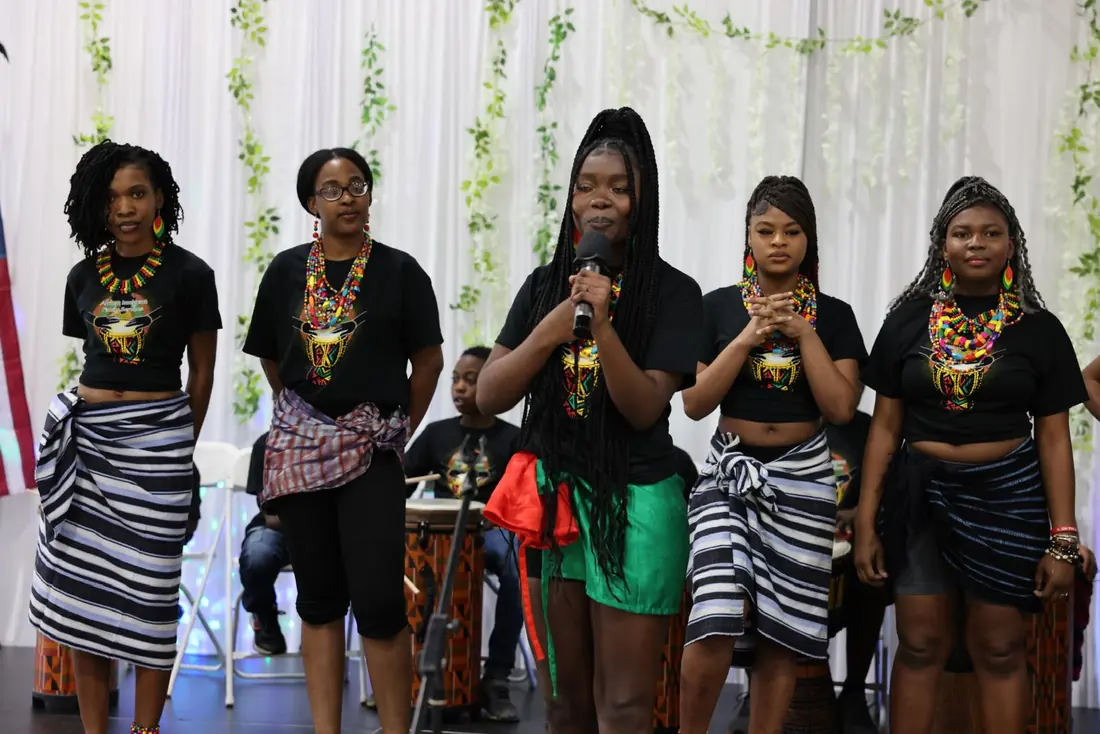 An Academy performer speaks into a microphone beside fellow performers in beaded necklaces and woven wraps, with drummers seated behind them