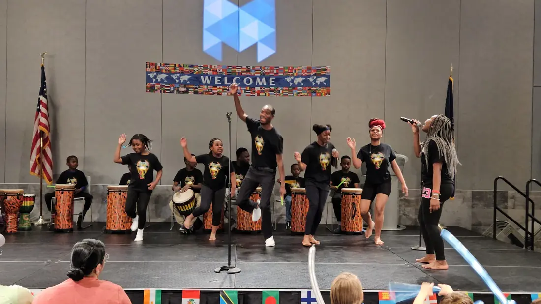 AIFS Youth Drumming Academy dancers leap and a vocalist sings while youth drummers play behind them on a stage under a Welcome banner of world flags