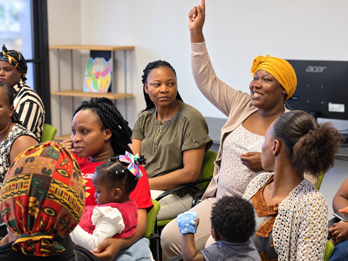 Mothers and young children at an AIFS family session, one woman in a yellow headwrap smiling and raising her hand to speak